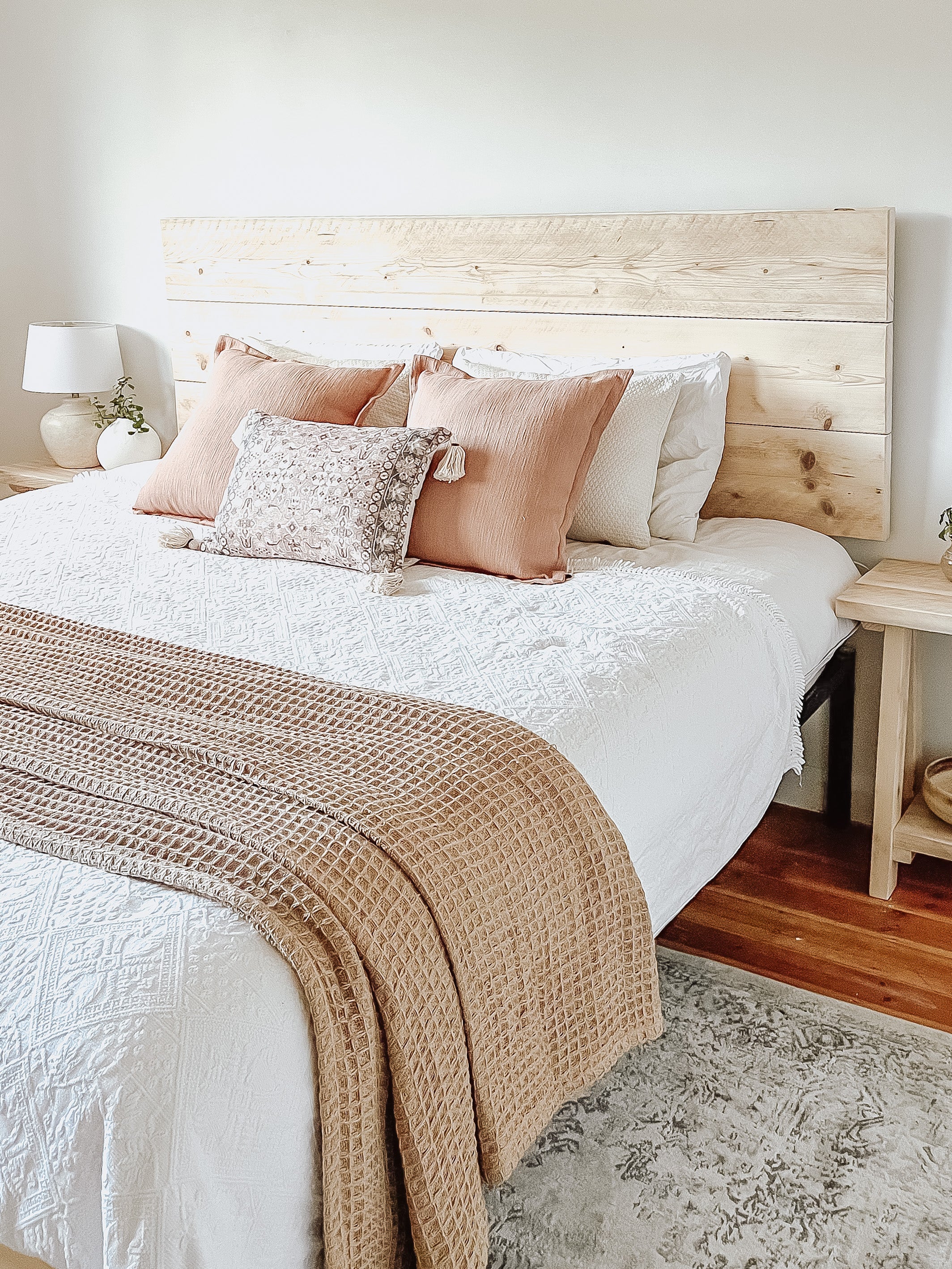 Rustic wooden Headboard with white sheets and  pink cushions, two rustic bedside tables with white farmhouse concrete style lamps