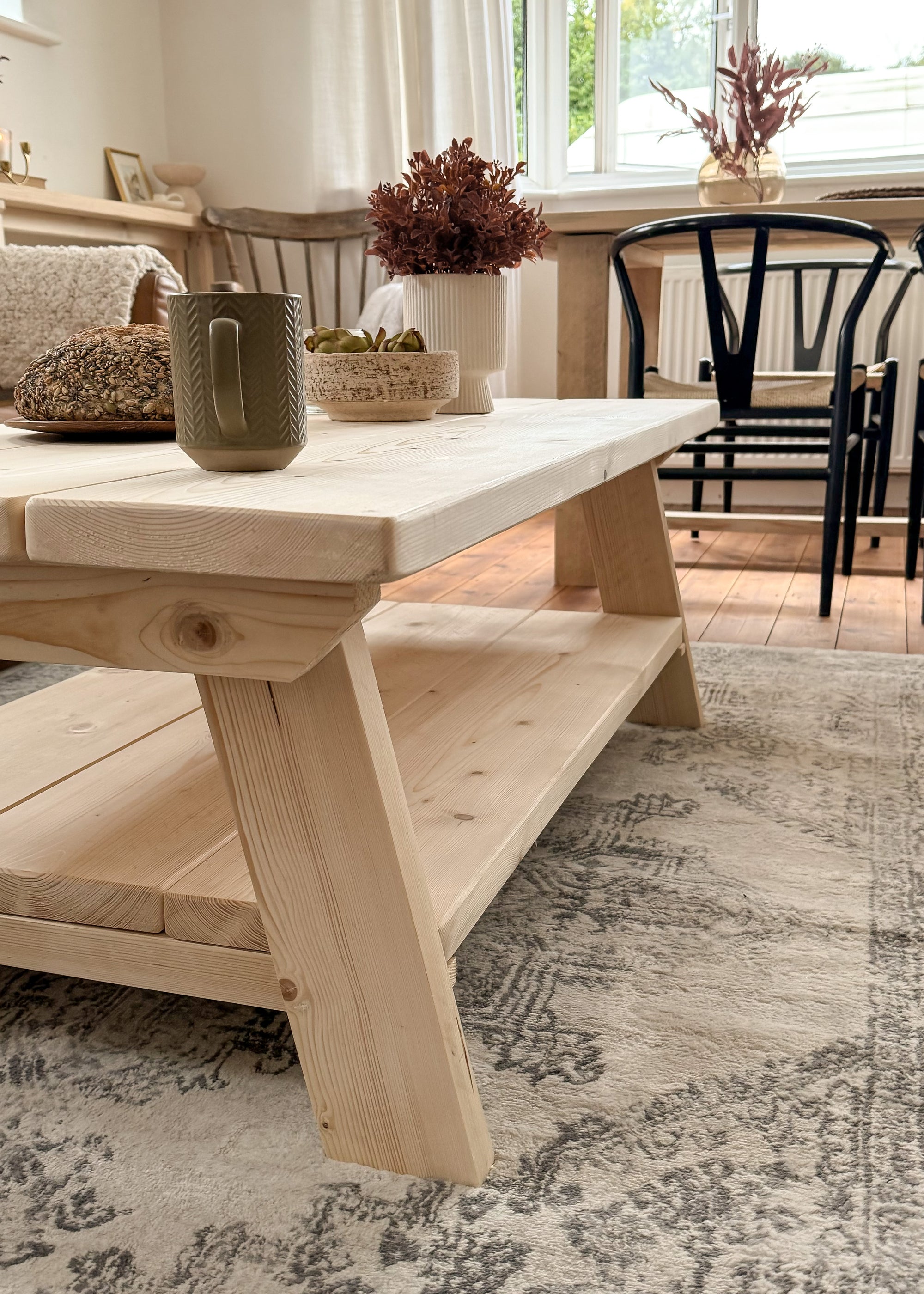 Rustic reclaimed wooden coffee table with a brown leather sofa in a stylish white-walled living room. The coffee table is adorned with a white vase of autumnal flowers, a loaf of fresh bread, and a cup. A light-colored area rug complements the cozy, inviting space.
