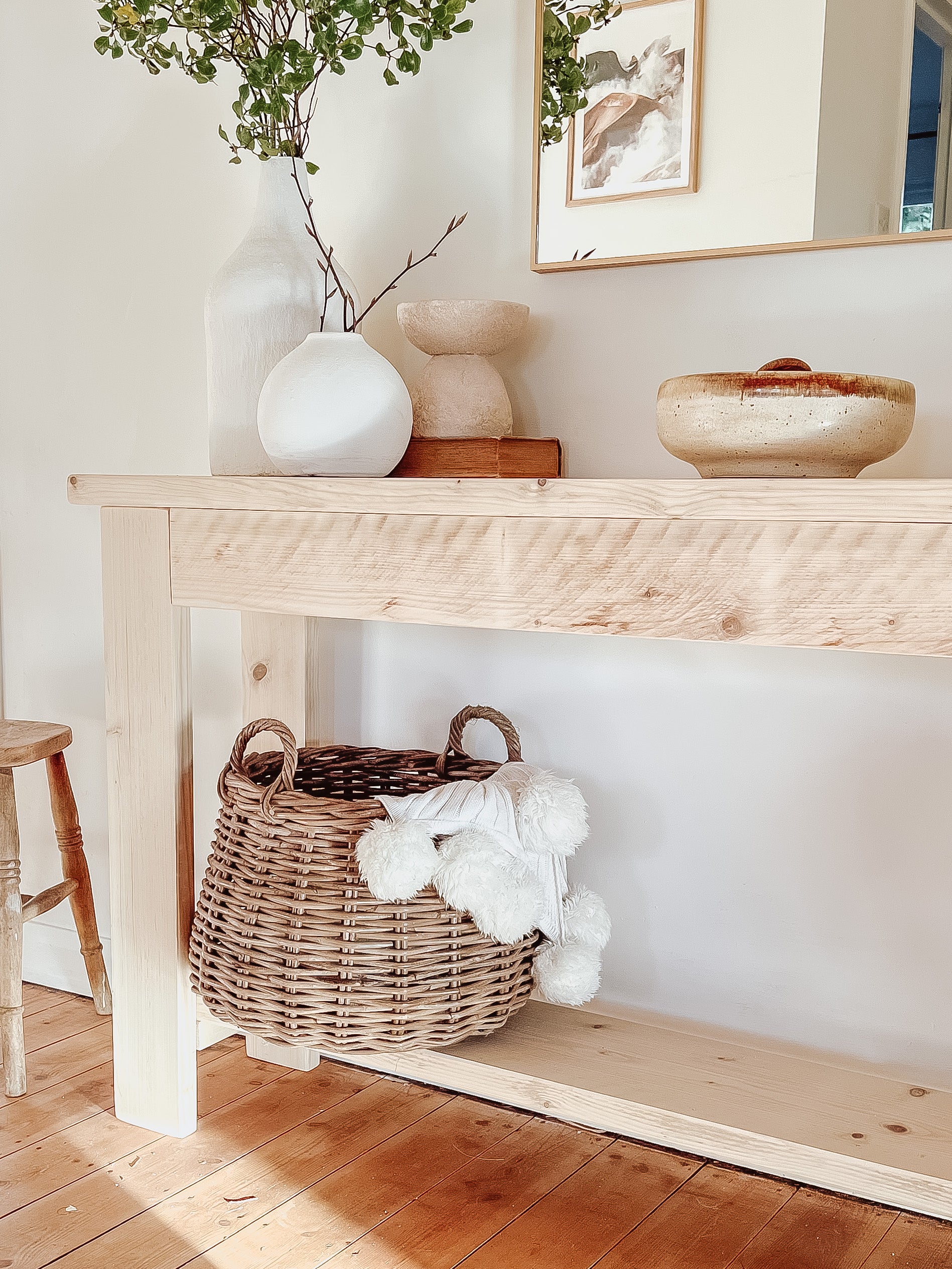 Rustic wood console table with vase and farmhouse lamp on a white background. Arched golden mirror above. Wicker basket holding a blanket below on the shelf of the console table. Wooden floors, a wooden stool sits next to the console entry way bench