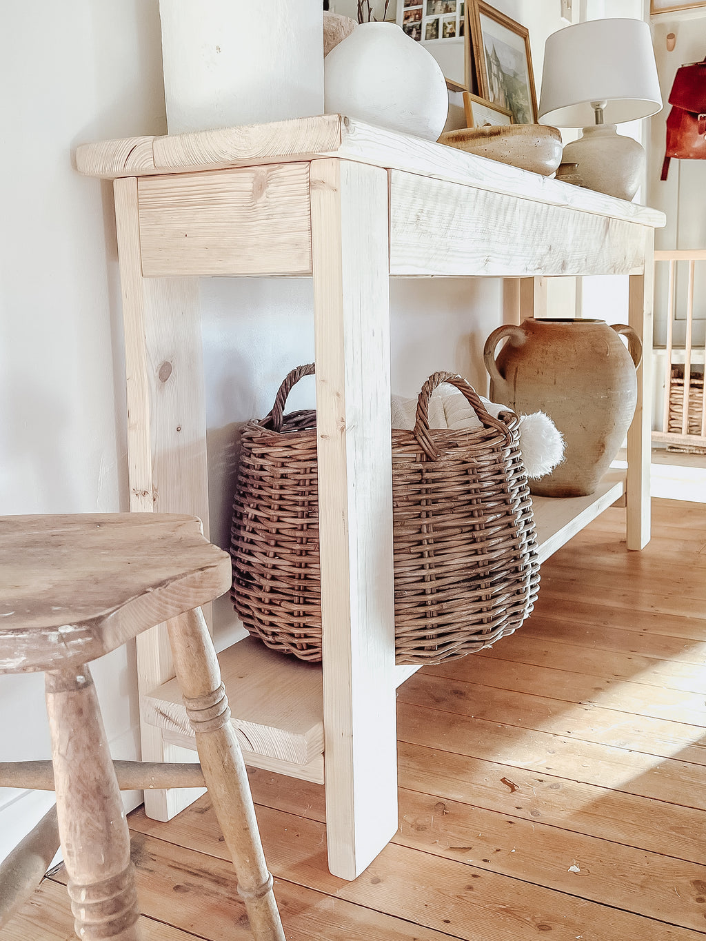 Rustic wood console table with vase and farmhouse lamp on a white background. Arched golden mirror above. Wicker basket holding a blanket below on the shelf of the console table. Wooden floors, a wooden stool sits next to the console entry way bench