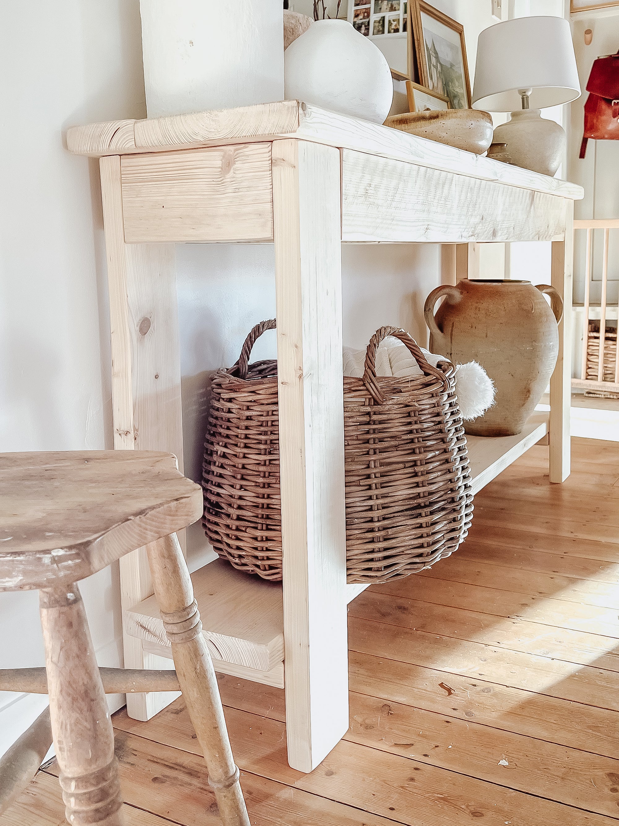 Rustic wood console table with vase and farmhouse lamp on a white background. Arched golden mirror above. Wicker basket holding a blanket below on the shelf of the console table. Wooden floors, a wooden stool sits next to the console entry way bench