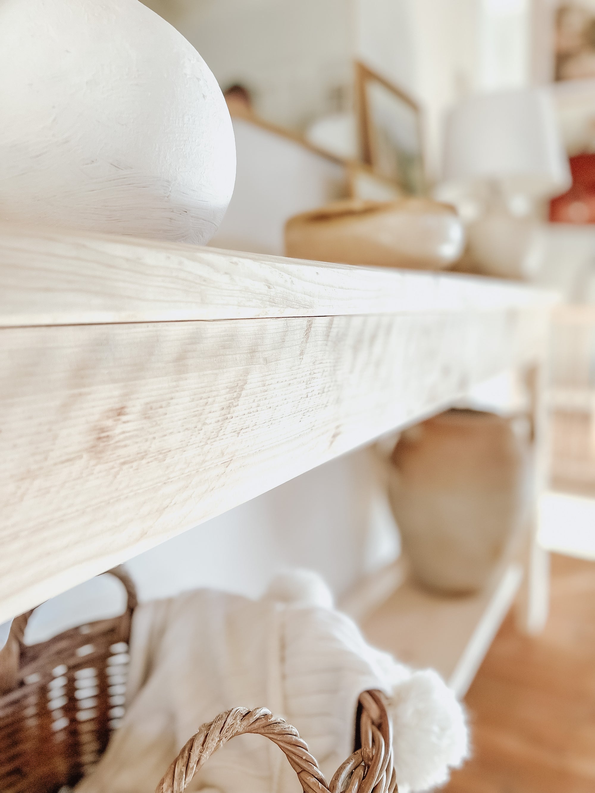 Rustic wood console table with vase and farmhouse lamp on a white background. Arched golden mirror above. Wicker basket holding a blanket below on the shelf of the console table. Wooden floors, a wooden stool sits next to the console entry way bench
