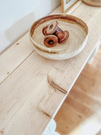 Rustic wood console table with vase and farmhouse lamp on a white background. Arched golden mirror above. Wicker basket holding a blanket below on the shelf of the console table. Wooden floors, a wooden stool sits next to the console entry way bench
