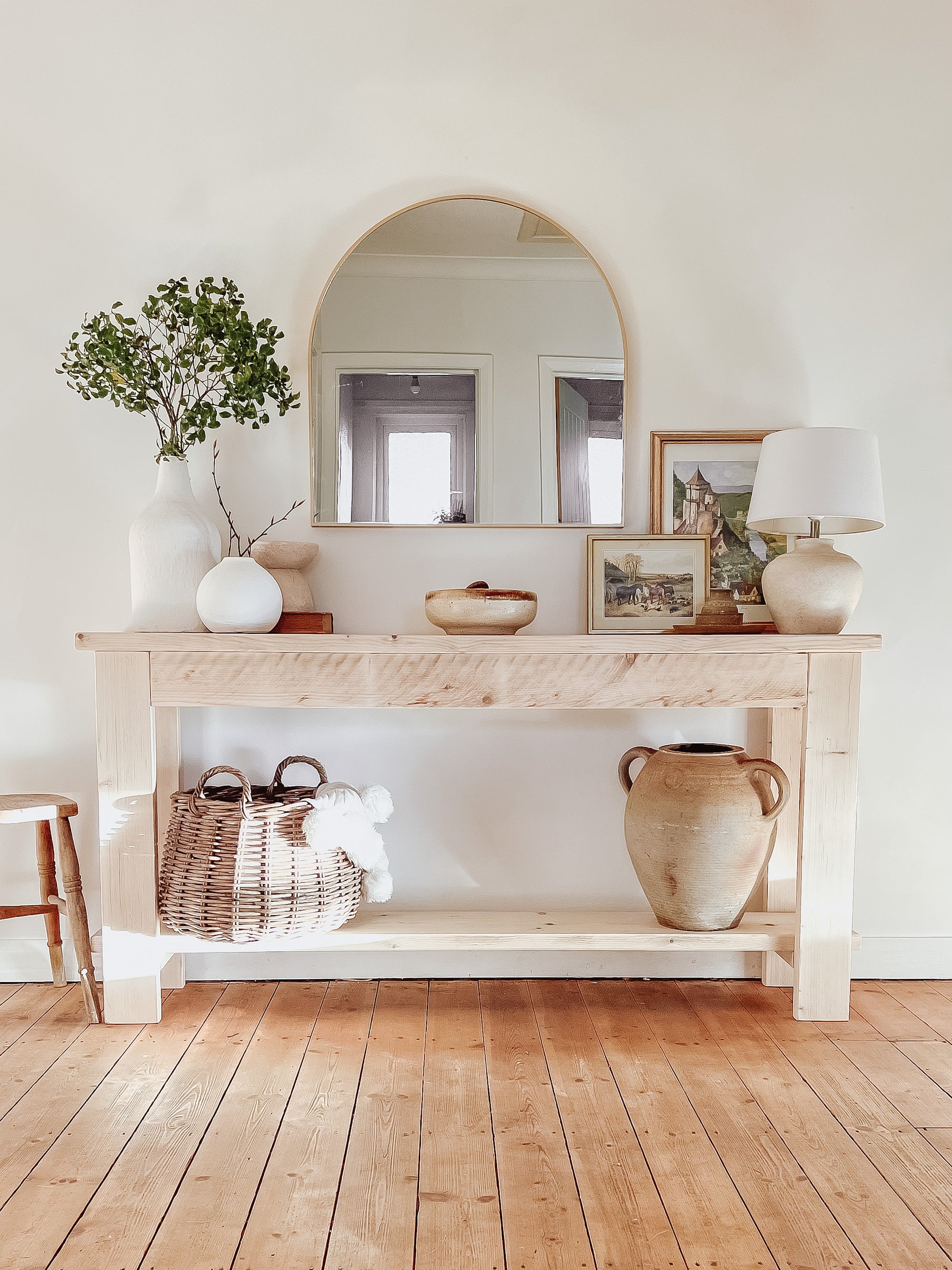 Rustic wood console table with vase and farmhouse lamp on a white background. Arched golden mirror above. Wicker basket holding a blanket below on the shelf of the console table. Wooden floors, a wooden stool sits next to the console entry way bench