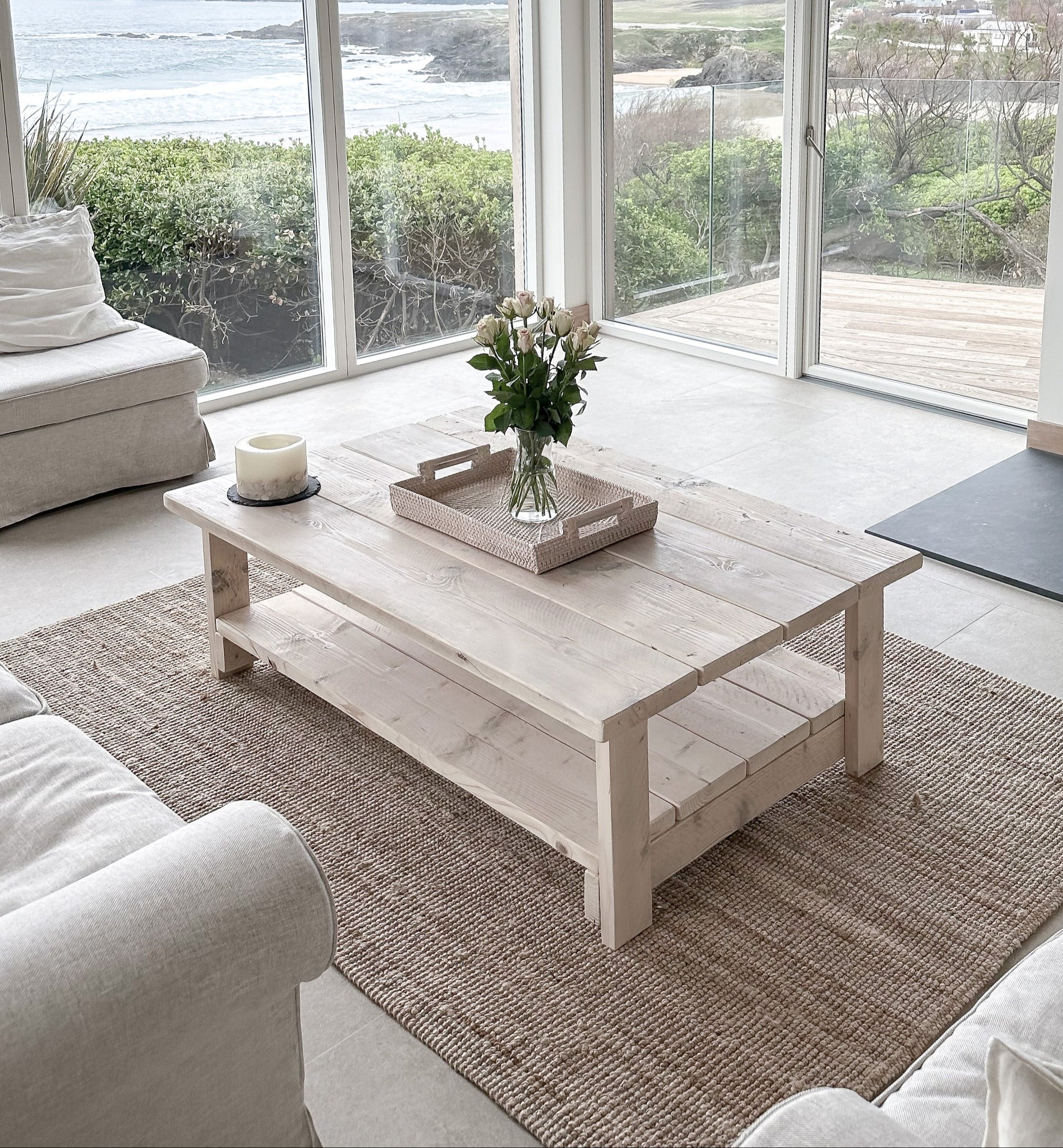 Whitewashed coffee table in a Modern living room with large windows, white furniture, and a fireplace.