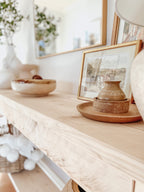 Rustic wood console table with vase and farmhouse lamp on a white background. Arched golden mirror above. Wicker basket holding a blanket below on the shelf of the console table. Wooden floors, a wooden stool sits next to the console entry way bench