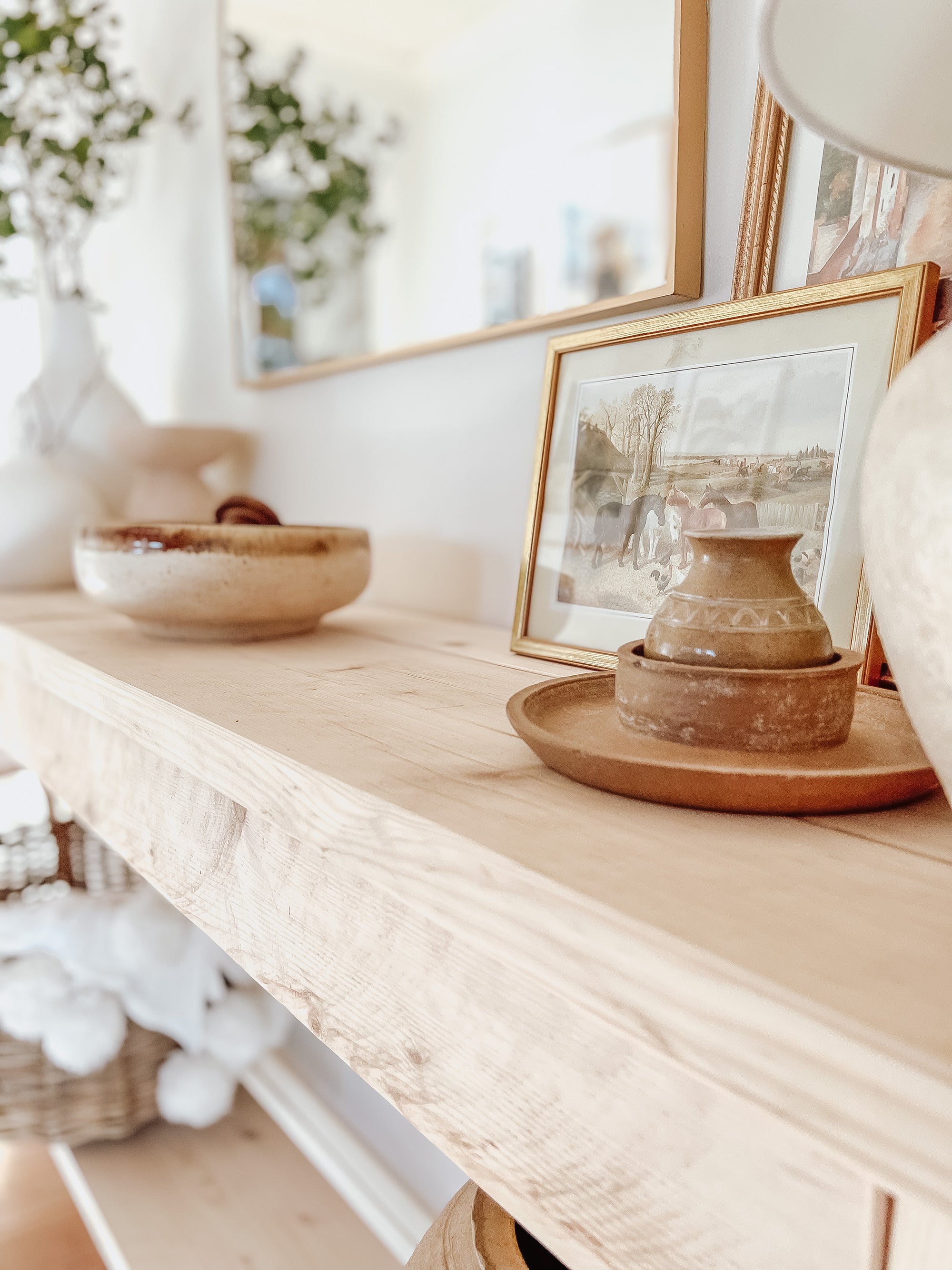 Rustic wood console table with vase and farmhouse lamp on a white background. Arched golden mirror above. Wicker basket holding a blanket below on the shelf of the console table. Wooden floors, a wooden stool sits next to the console entry way bench