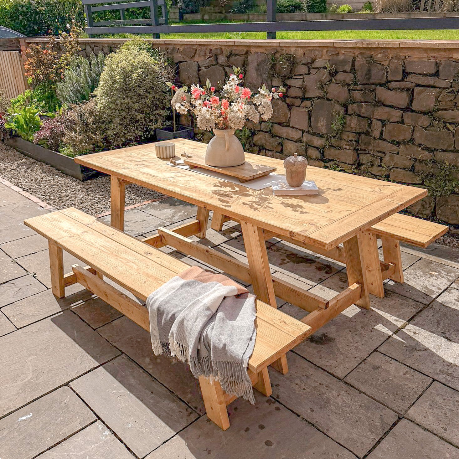 Wooden outdoor dining table with benches on a patio, set with a blanket and flowers.