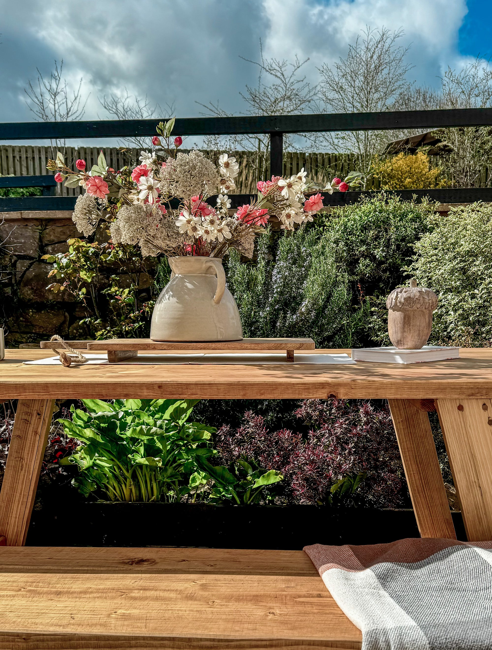 Wooden outdoor table with a vase of flowers and a potted plant on a garden background
