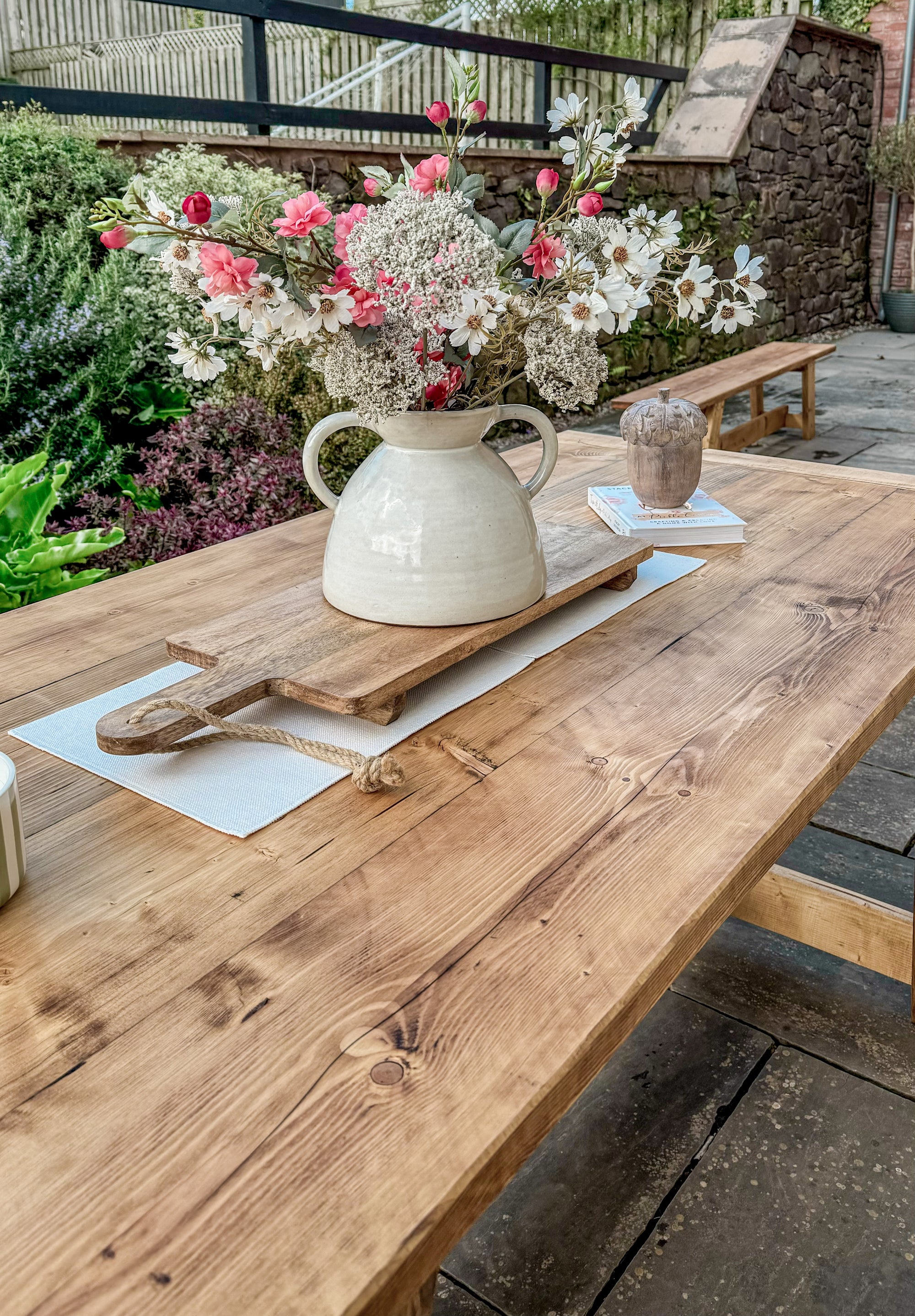 Wooden table with a vase of flowers and a cutting board outdoors.