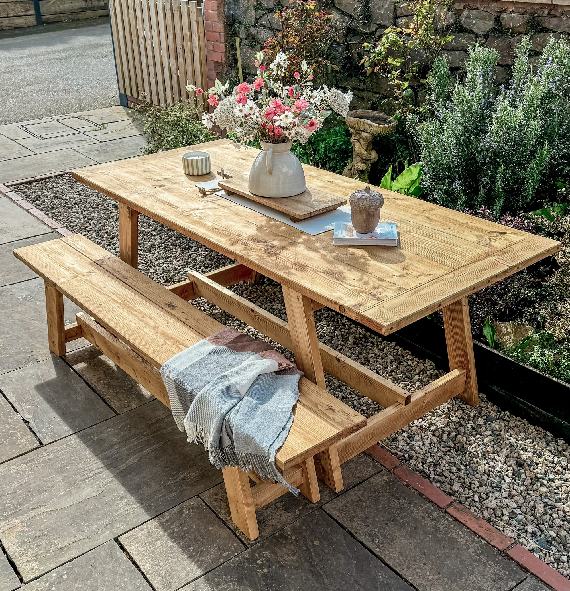 Wooden outdoor table with benches and a vase of flowers in a garden setting.