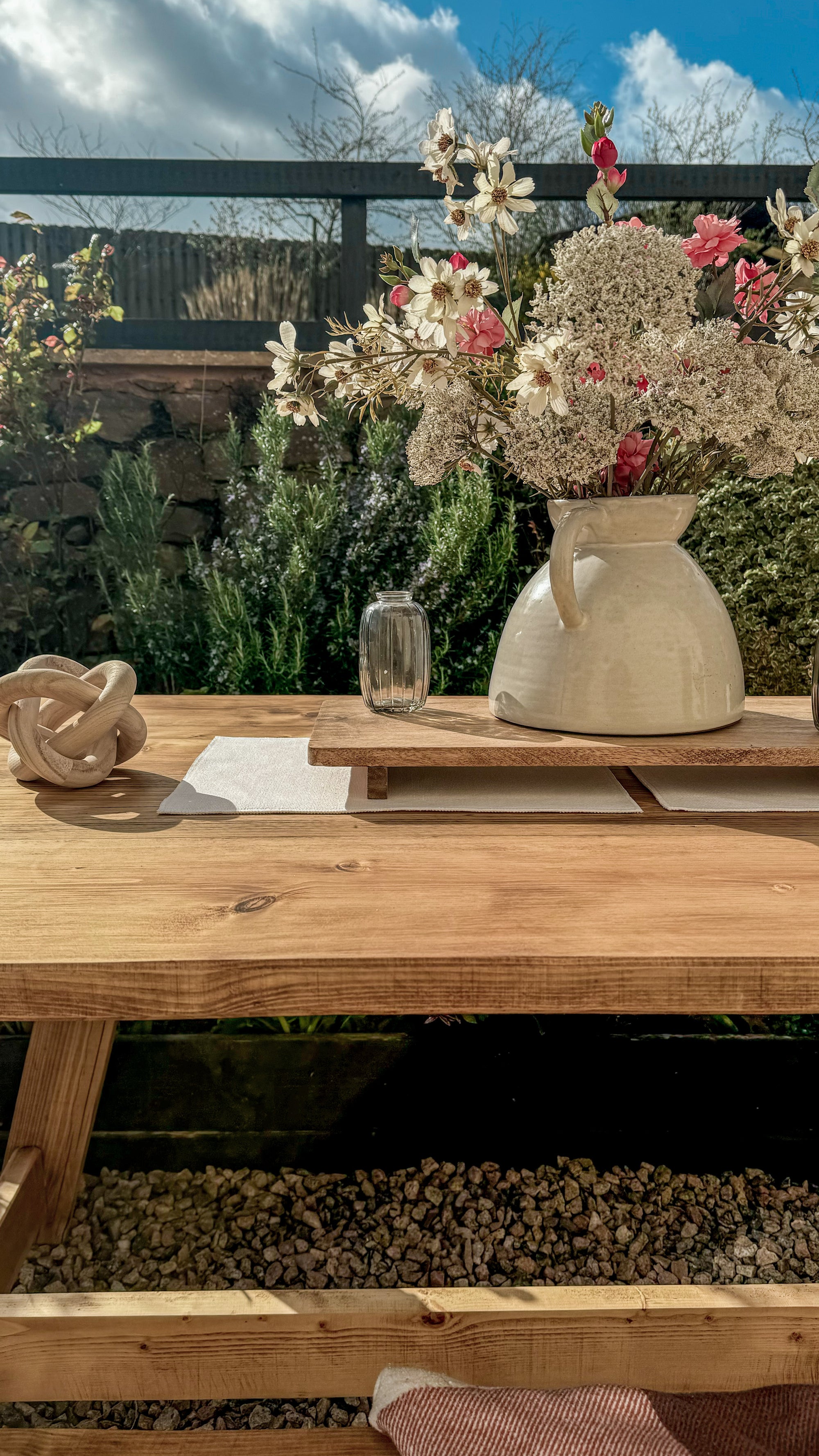 Wooden table with decorative items including a vase of flowers and a teapot in an outdoor setting.