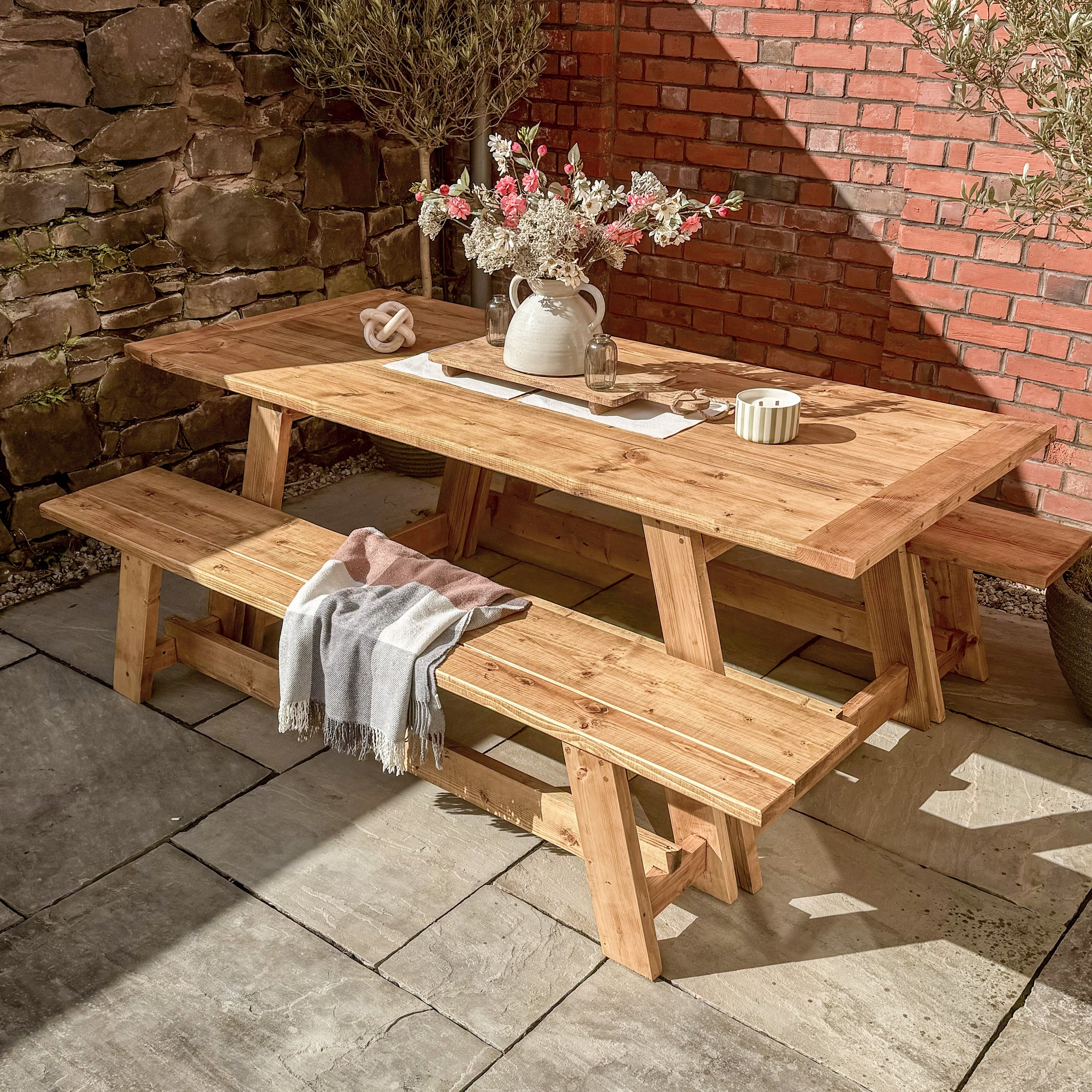 Wooden picnic table with benches in a garden setting with a stone wall and brick wall in the background.