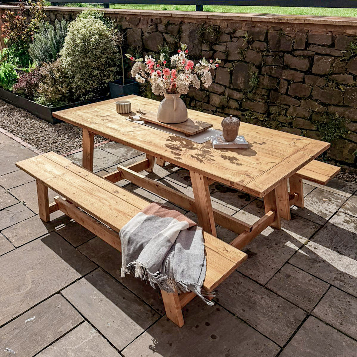Wooden outdoor table with benches on a patio, featuring a vase of flowers and decorative items.