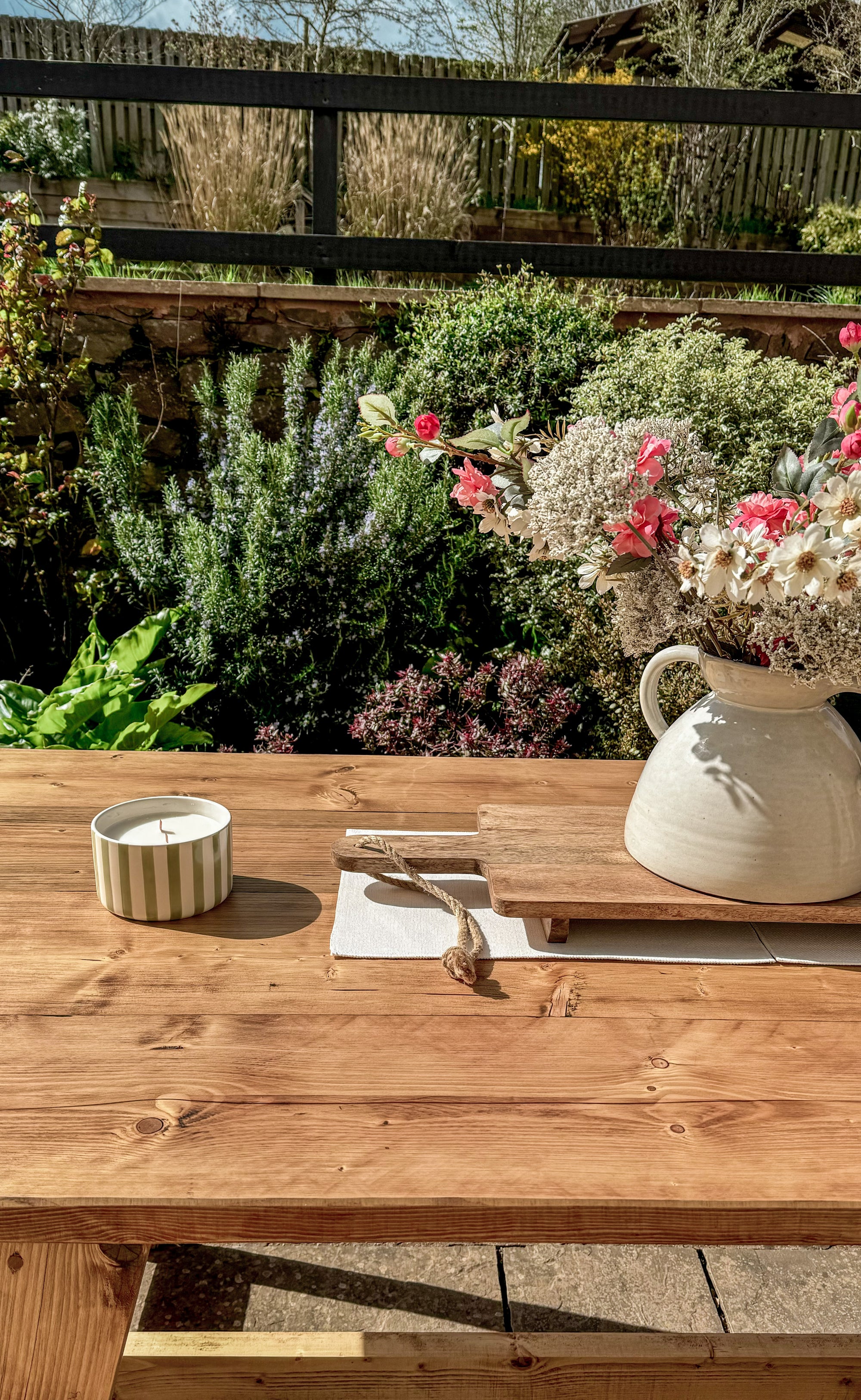 Wooden outdoor table with a vase of flowers, candle, and book in a garden setting