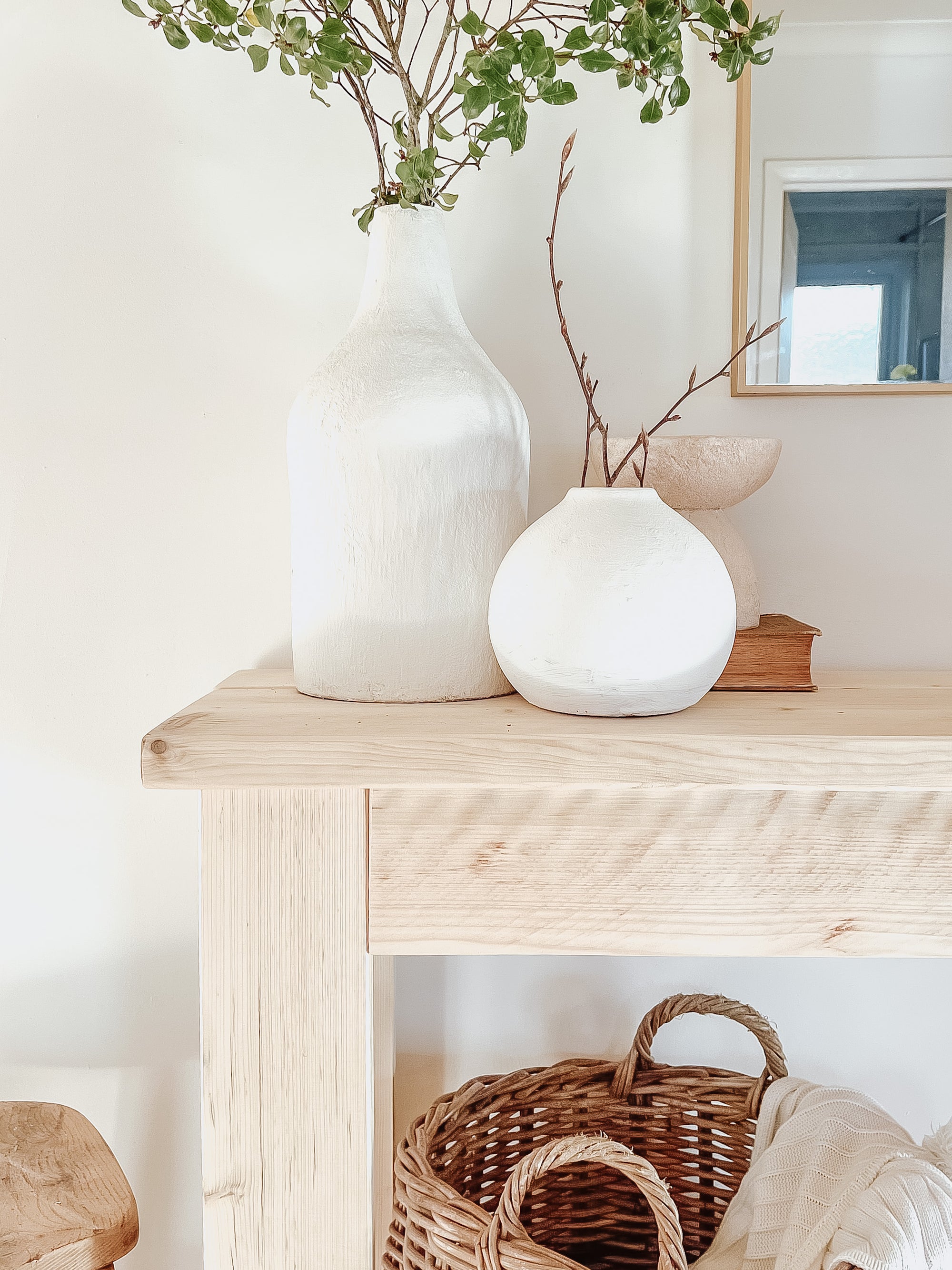 Rustic wood console table with vase and farmhouse lamp on a white background. Arched golden mirror above. Wicker basket holding a blanket below on the shelf of the console table. Wooden floors, a wooden stool sits next to the console entry way bench