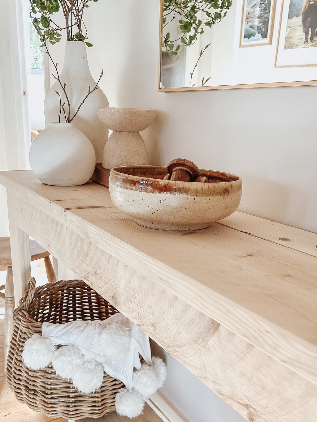 Rustic wood console table with vase and farmhouse lamp on a white background. Arched golden mirror above. Wicker basket holding a blanket below on the shelf of the console table. Wooden floors, a wooden stool sits next to the console entry way bench