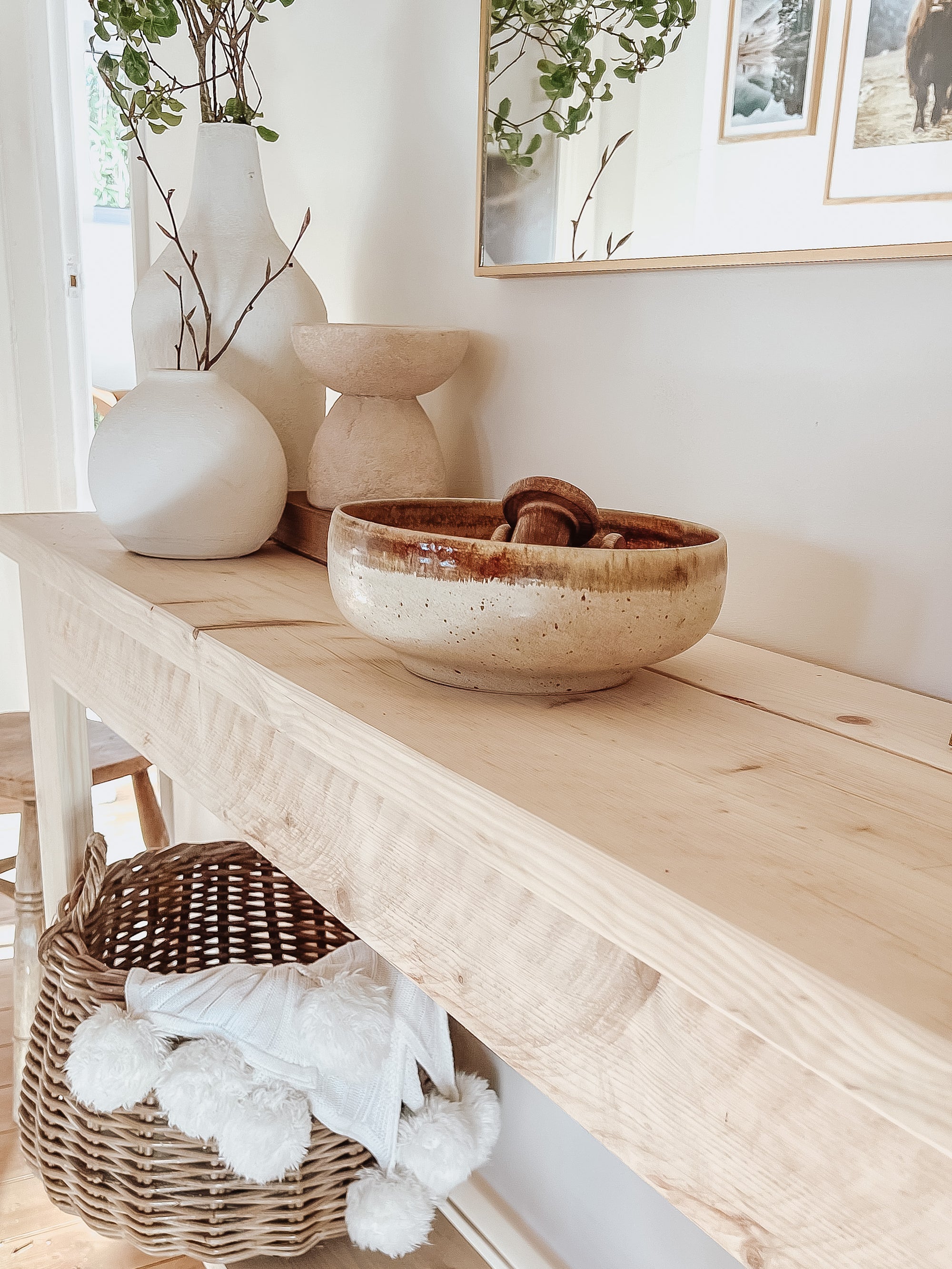 Rustic wood console table with vase and farmhouse lamp on a white background. Arched golden mirror above. Wicker basket holding a blanket below on the shelf of the console table. Wooden floors, a wooden stool sits next to the console entry way bench