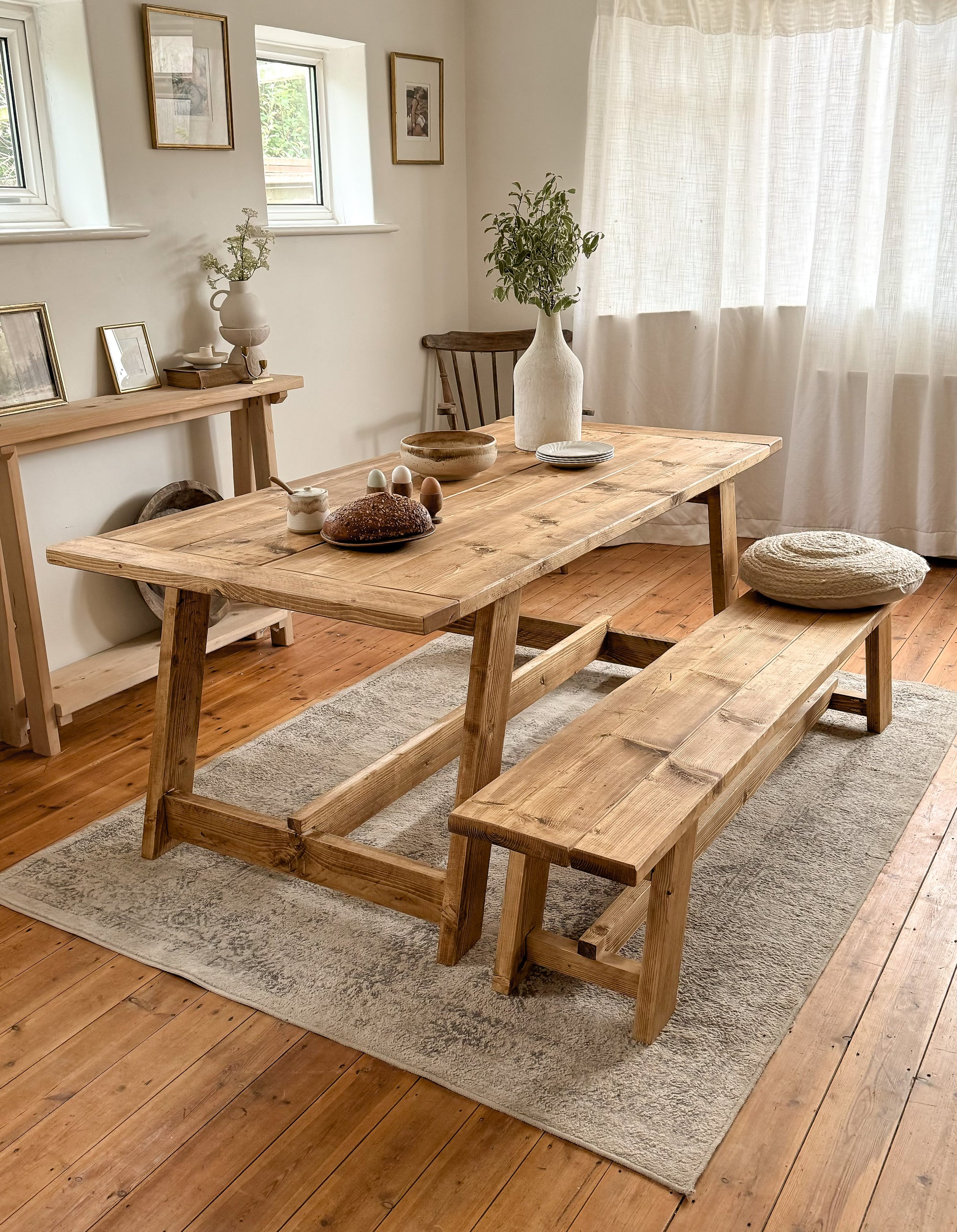 Rustic dining table and bench, sitting on a light coloured rug with white walls. It shows the ends of the table, and the end of the bench. The wood is antique oak. 