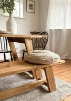 A close up of the rustic dining table bench, with a cushion and a chair in the background. The finish of the bench is antique oak.