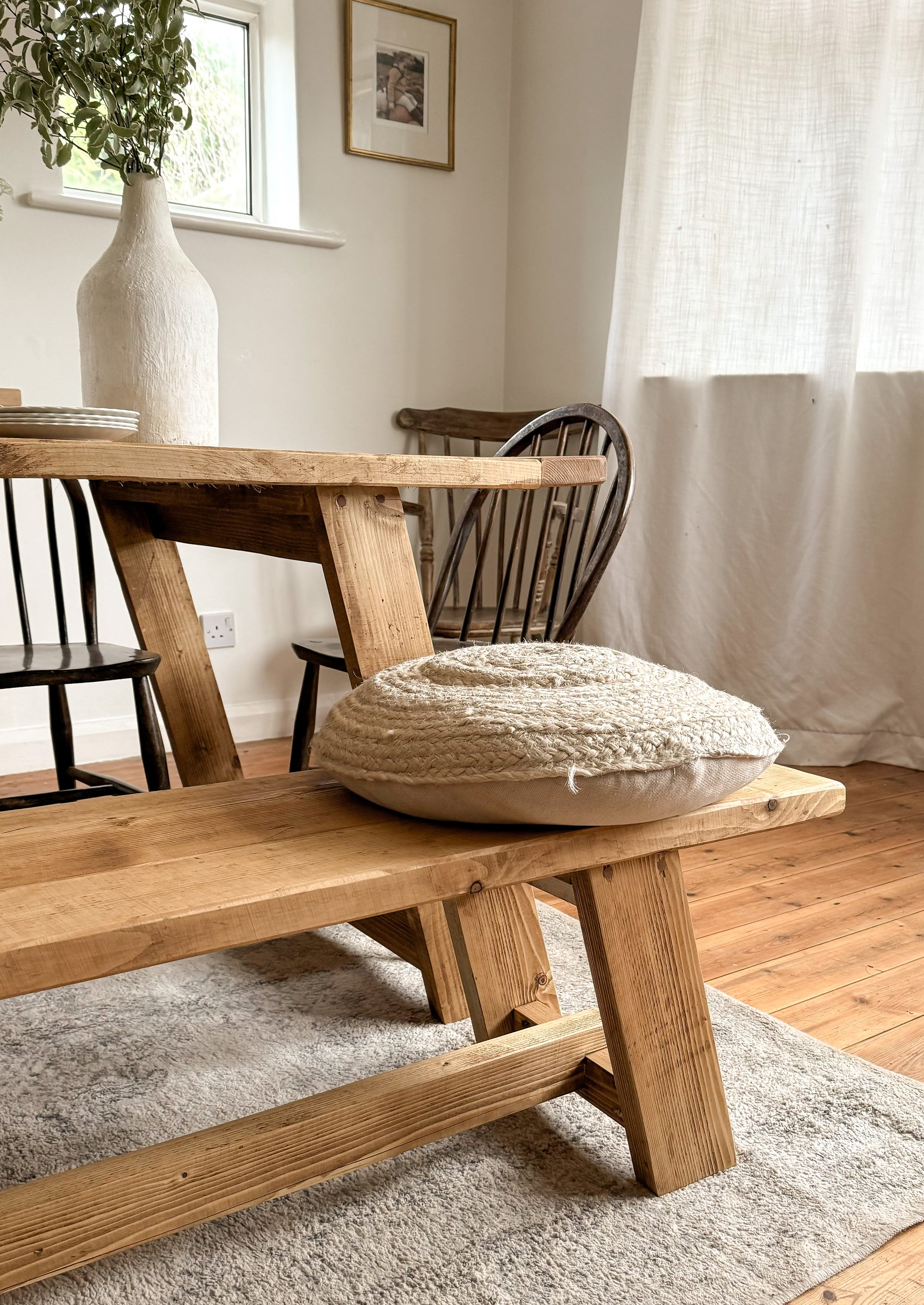 A close up of the rustic dining table bench, with a cushion and a chair in the background. The finish of the bench is antique oak.