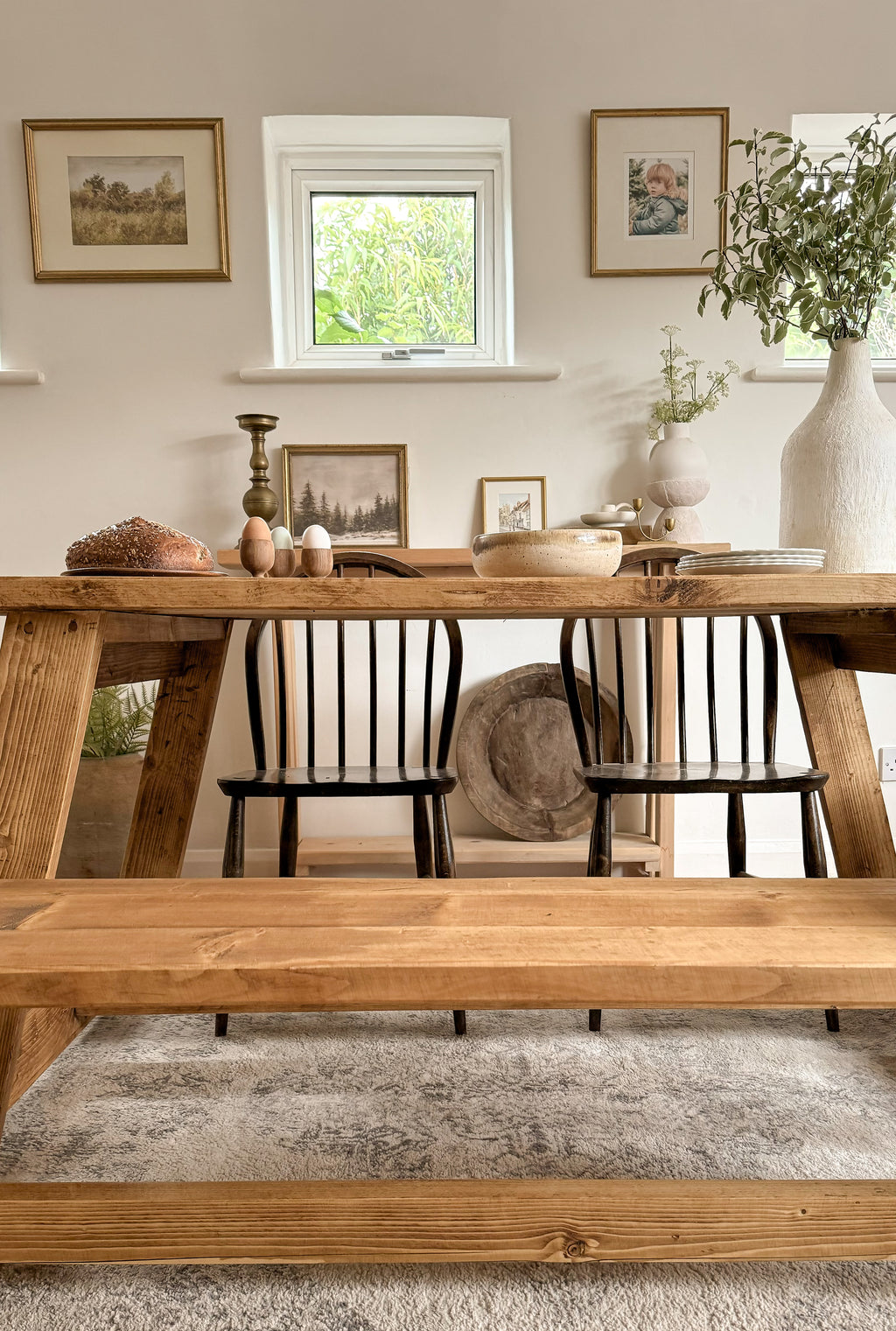Lifestyle image showing the farmhouse rustic dining table in a dining room, with a bench, 2 chairs, and an antique dough bowl on the sideboard behind. There are gold pictures on the wall, a vase, eggs on the table and bread.