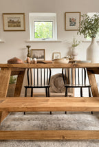Lifestyle image showing the farmhouse rustic dining table in a dining room, with a bench, 2 chairs, and an antique dough bowl on the sideboard behind. There are gold pictures on the wall, a vase, eggs on the table and bread.