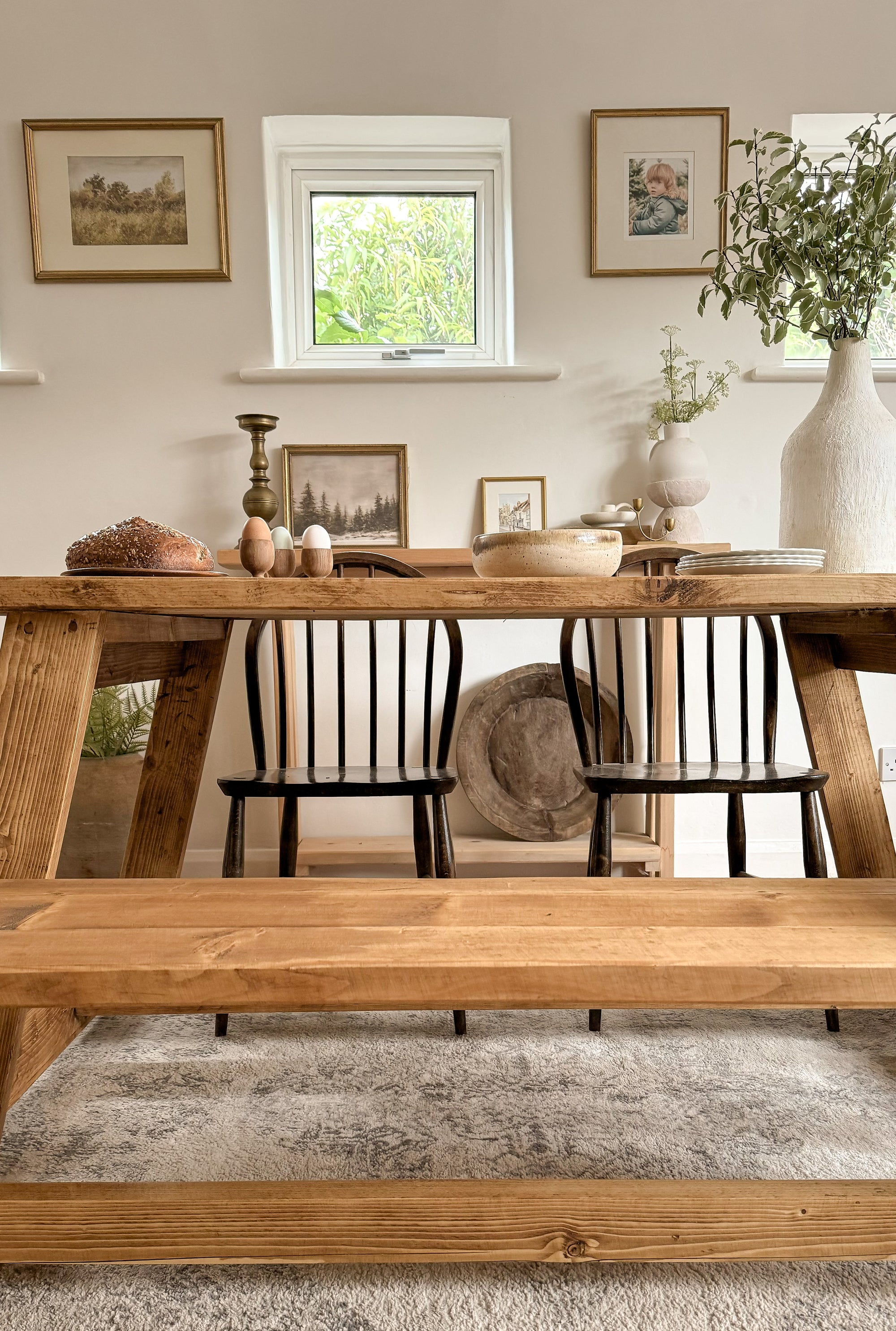 Lifestyle image showing the farmhouse rustic dining table in a dining room, with a bench, 2 chairs, and an antique dough bowl on the sideboard behind. There are gold pictures on the wall, a vase, eggs on the table and bread.